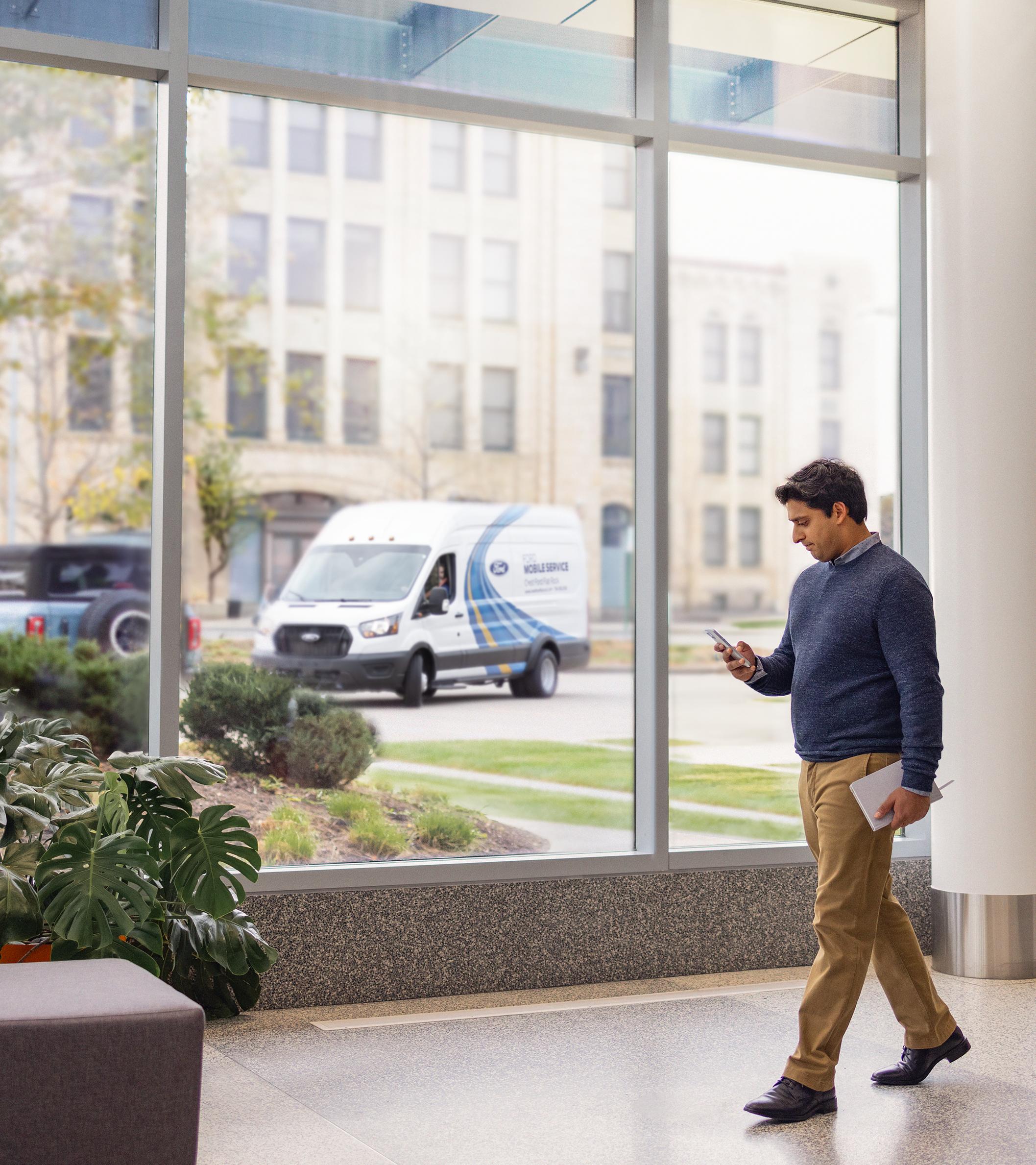 A person walks past a window inside a building while looking at their phone. Outside, a Mobile Service van is arriving
