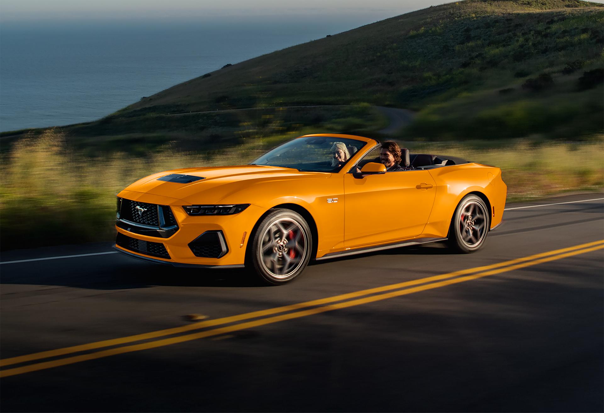 Two people in a 2026 Ford Mustang® GT Convertible in Orange Fury Metallic Tri-coat being driven on a coastal highway