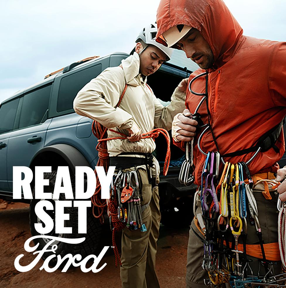 Two male rock climbers prepare for a climb beside a grey-blue Ford Bronco. White text “Ready Set Ford” is in the lower left corner.