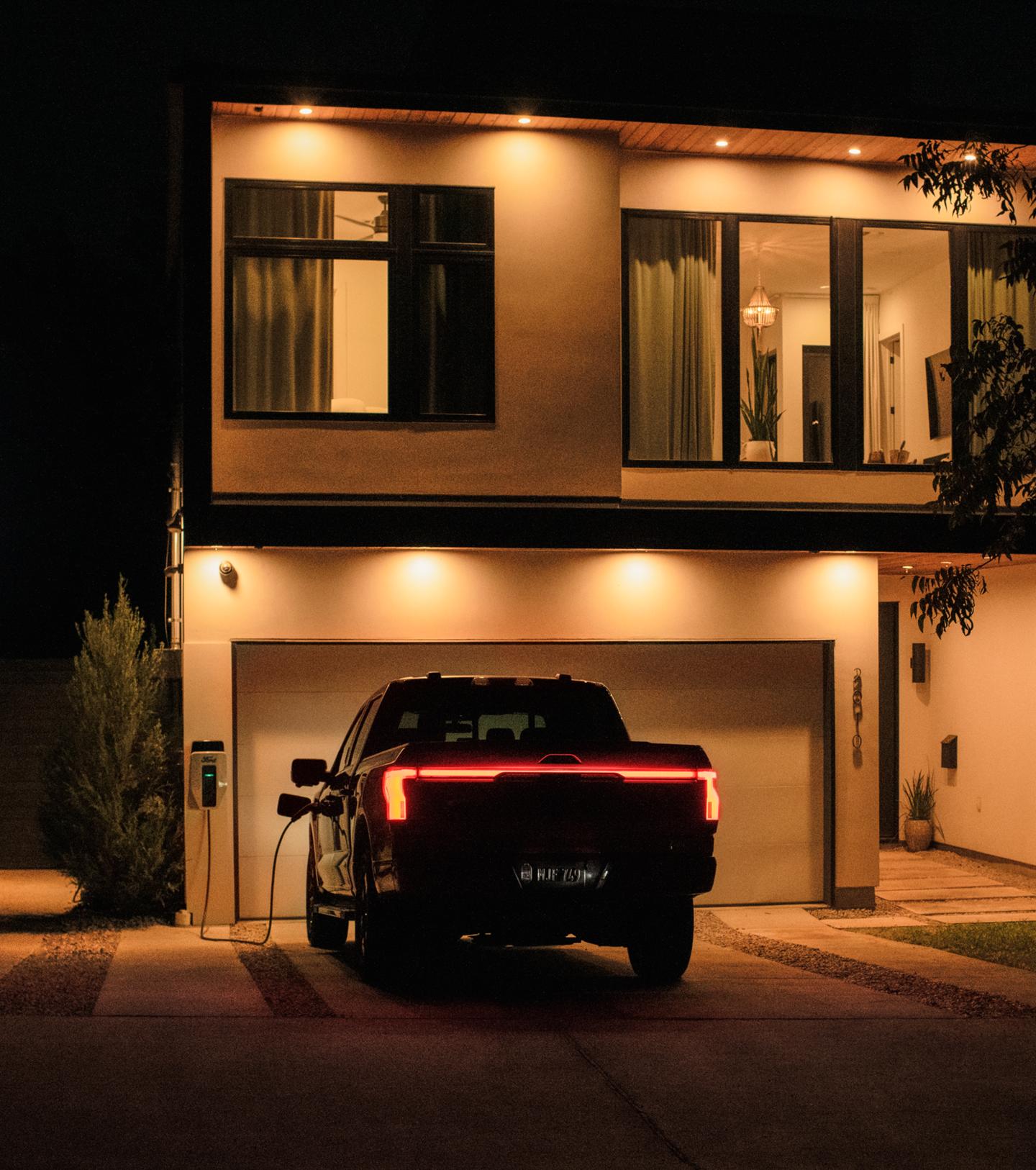 2025 Ford F-150® Lightning® pickup parked outside a home with a cord running from the truck to a wall-mounted charge station