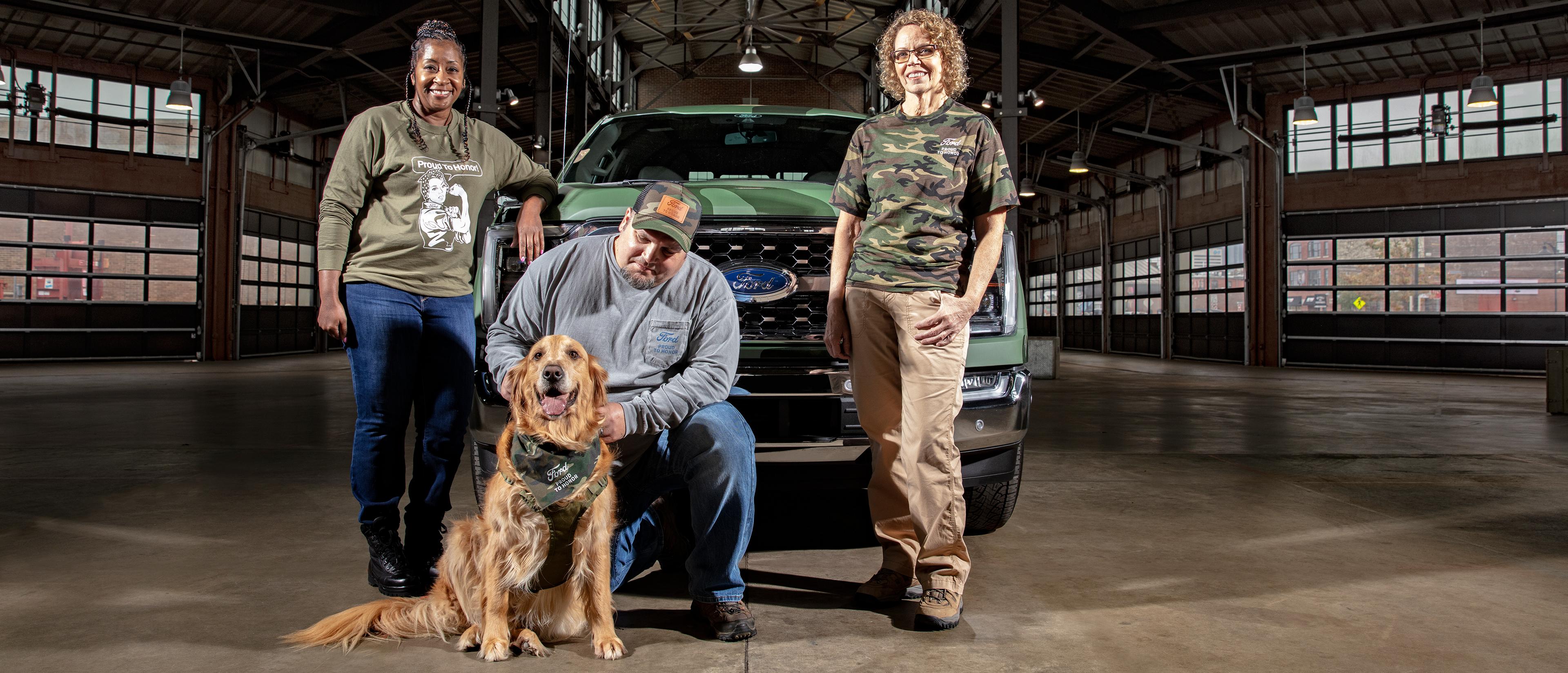 A group of people and a dog gather in front of a camoflauge-painted Ford truck in a warehouse