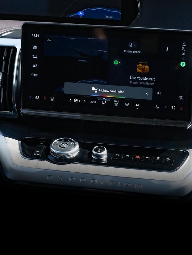 A close-up image of the center screen displaying a Google Voice Assistant prompt, asking “Hi, how can I help?”