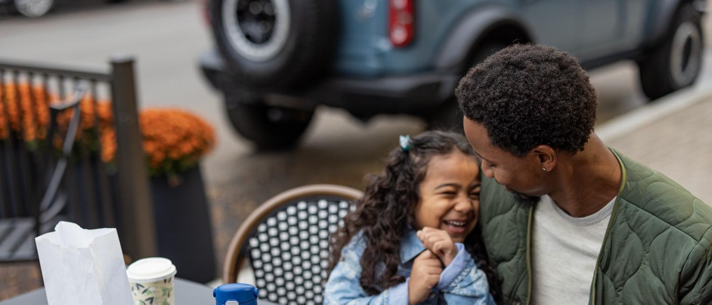 A father and daughter sit at an outdoor café with their Ford Bronco® parallel-parked in the background
