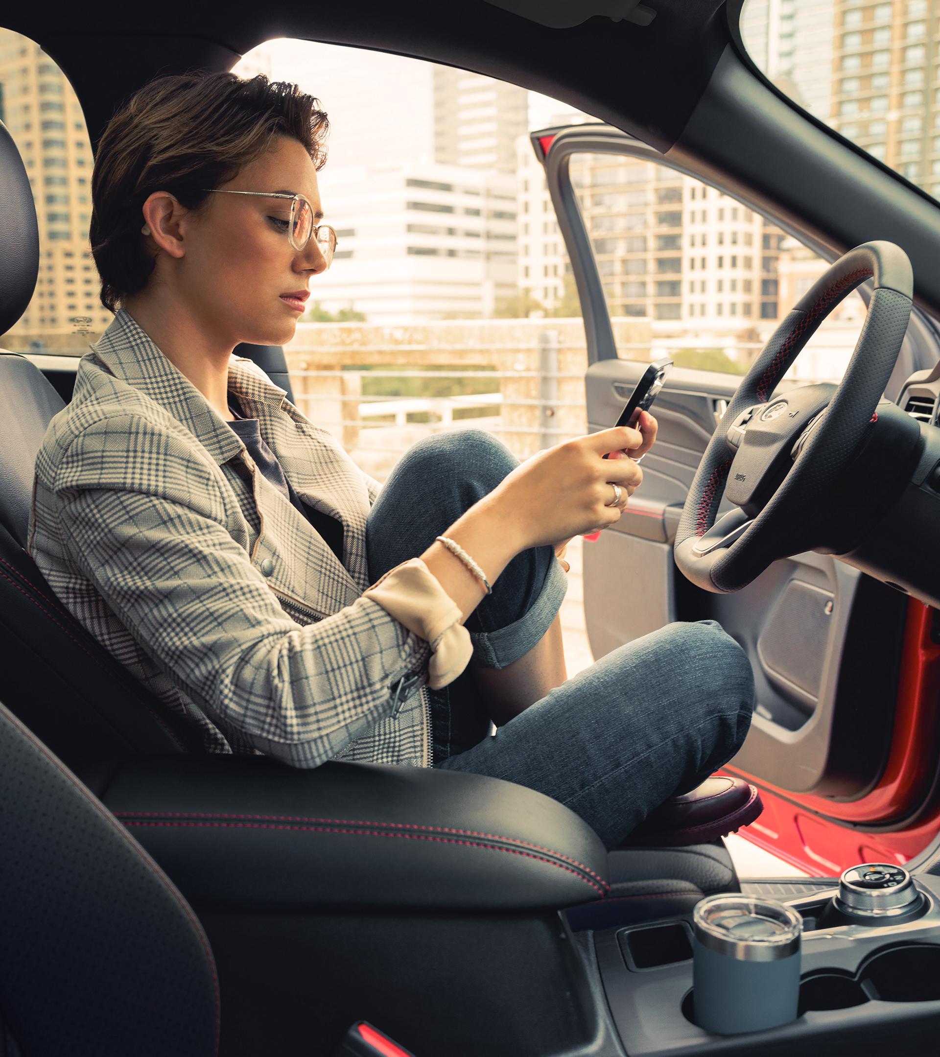 A Ford owner sits in the driver's seat glancing at their phone with the city skyline visible through the open door