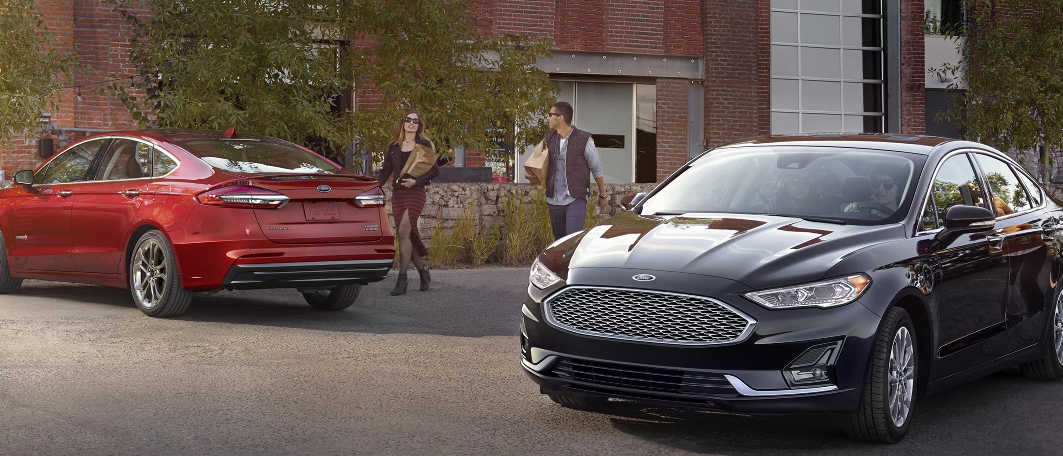 A couple of people carrying groceries approach a parked Ford Fusion vehicle while another Ford Fusion vehicle passes by