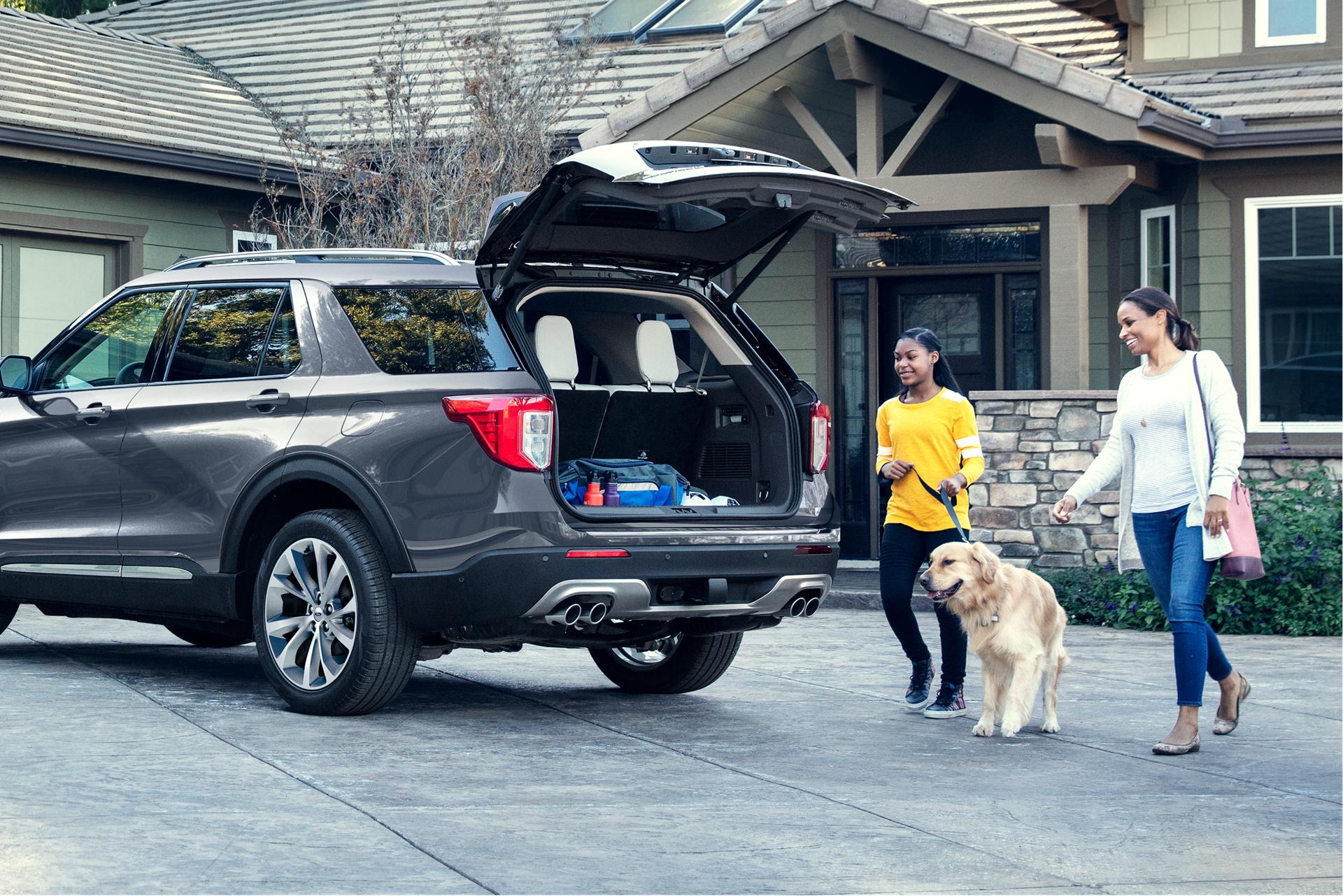 A couple of smiling people walk their dog over to the open liftgate of a Ford Explorer® SUV parked in the driveway