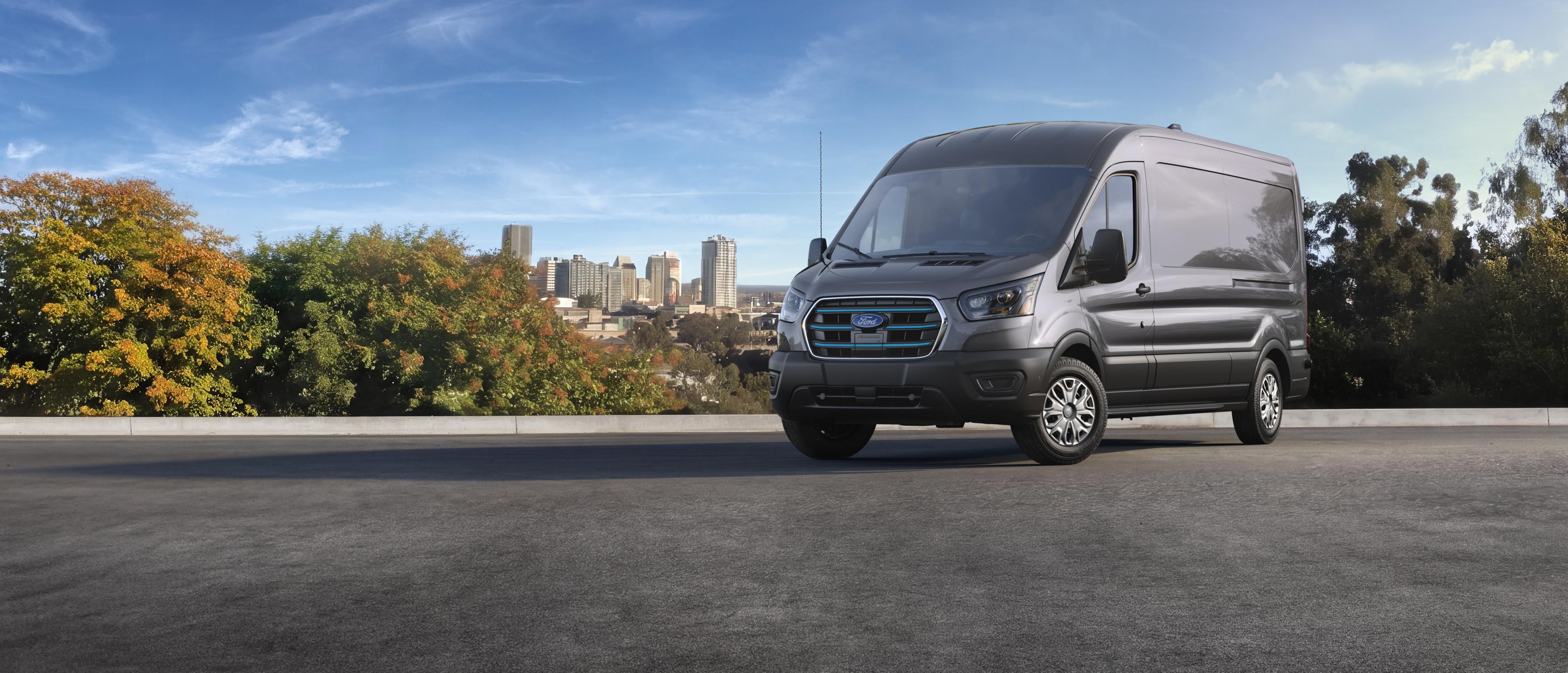 A gray 2027 Ford E-Transit™ Cargo Van parked, with a city skyline and trees in the background under a clear sky
