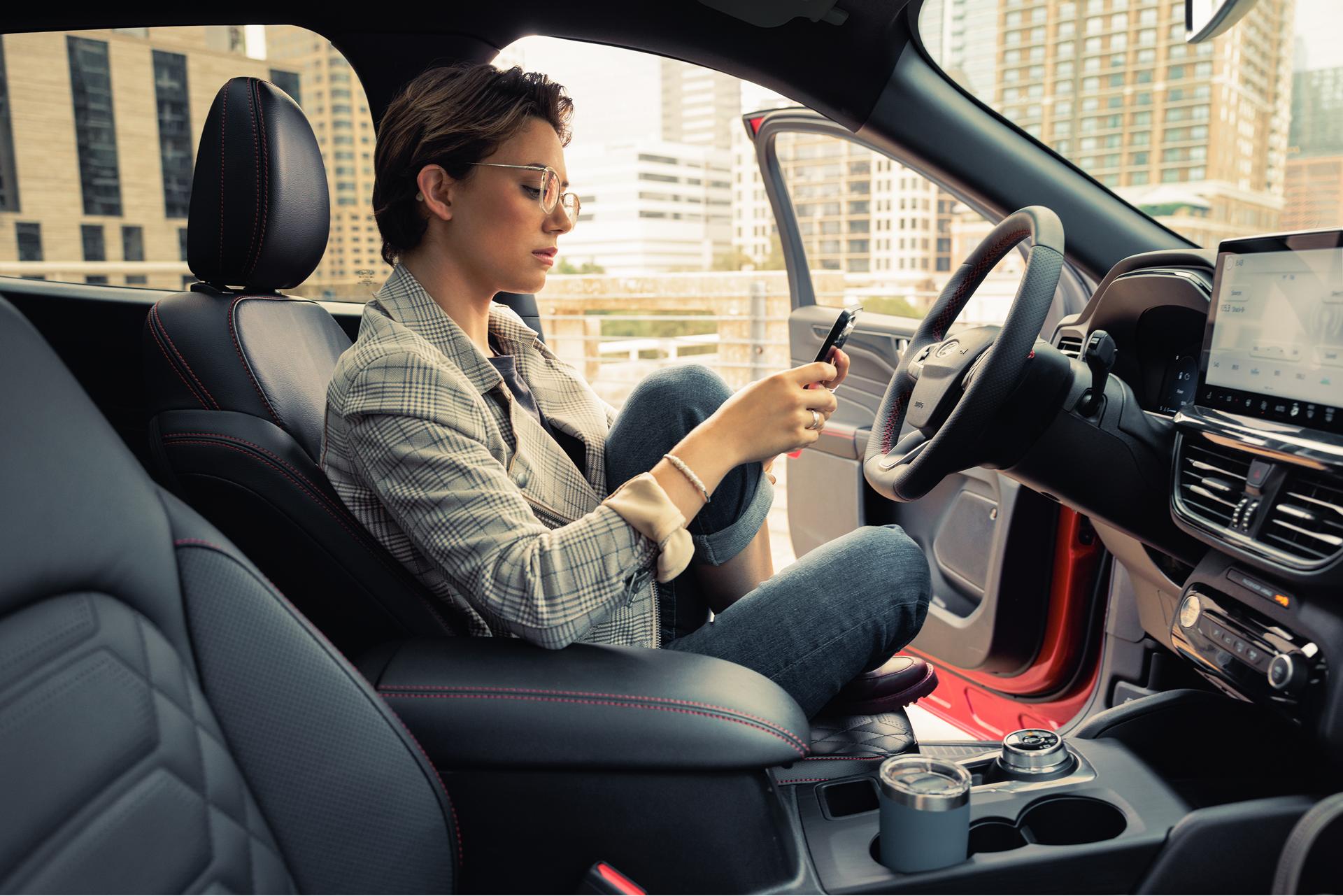 A woman checks her phone while sitting in the driver’s seat of her Ford vehicle with the door open