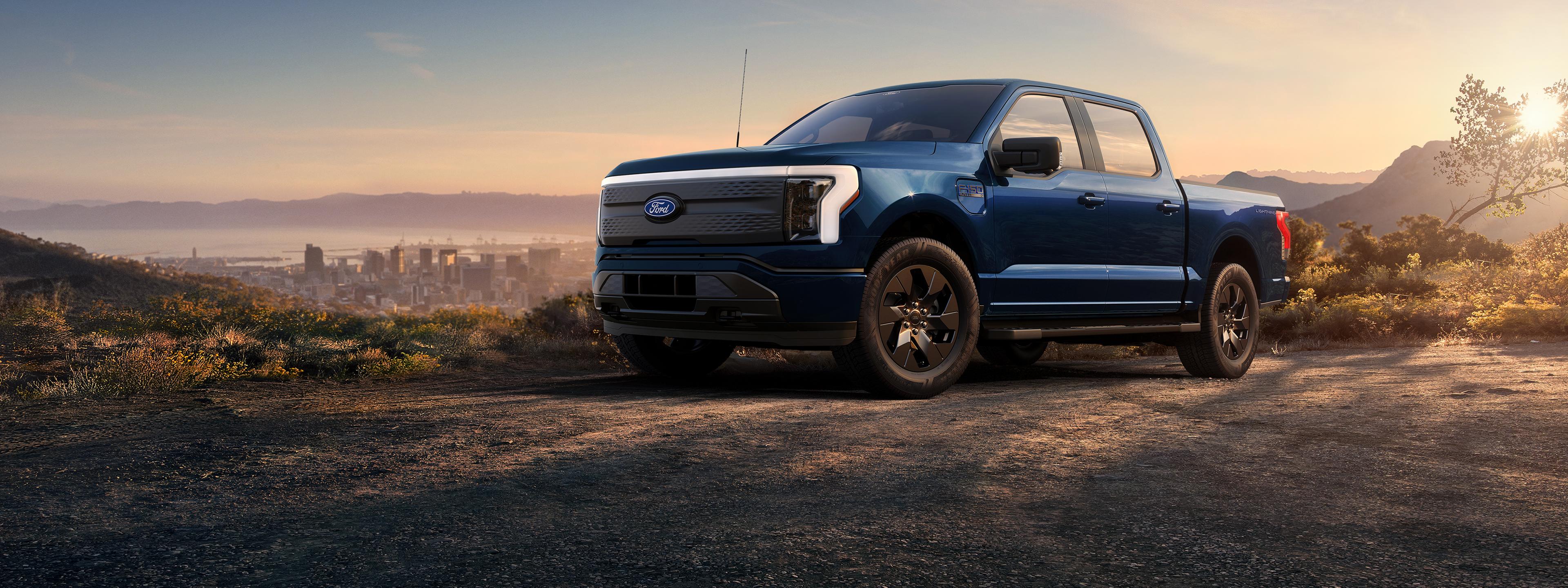 A blue 2025 Ford F-150® Lightning® truck parked in a  mountainous area. A city is in the background.