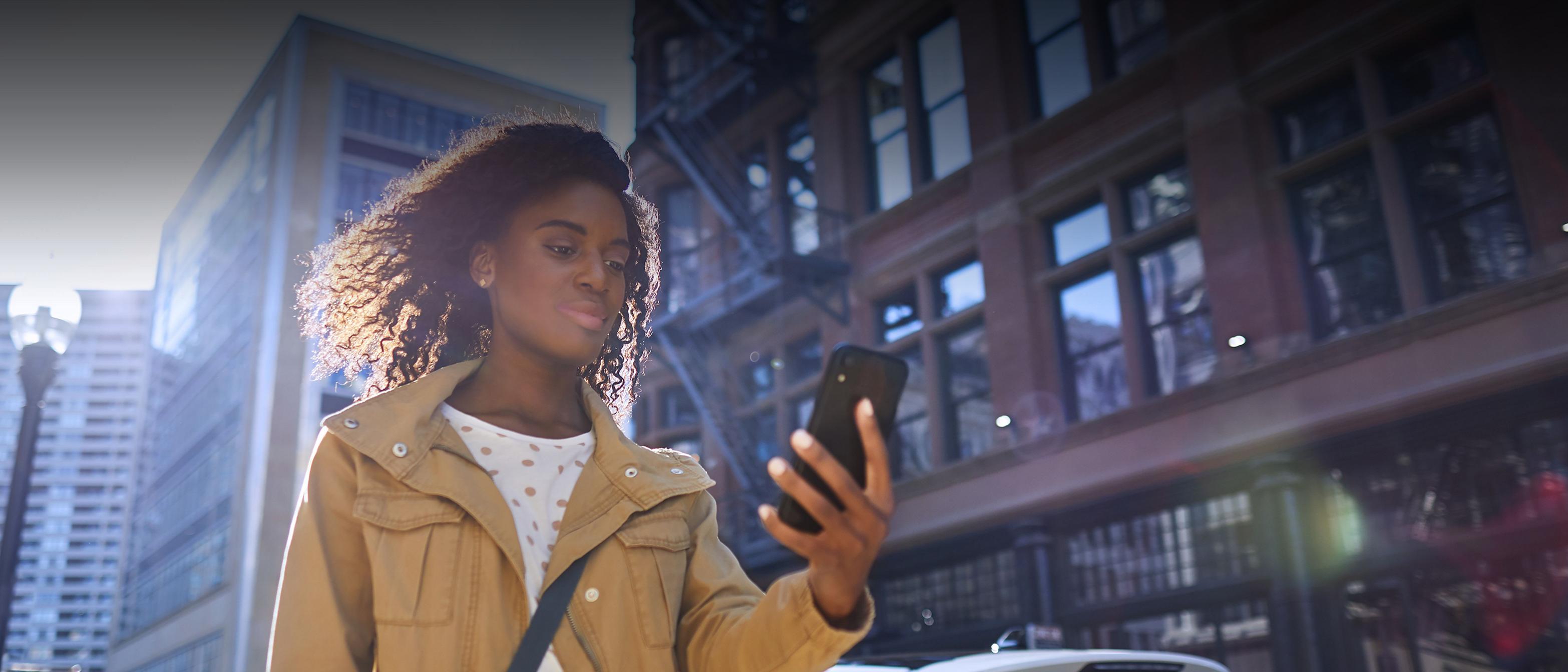 A woman walks on a sunny city street while looking at her smartphone