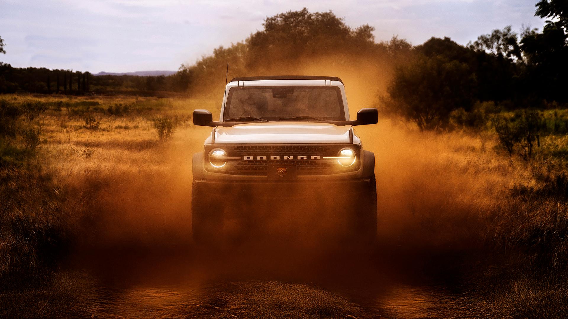 The front of a 2026 Ford Bronco® SUV, its headlights shining through a cloud of red dust