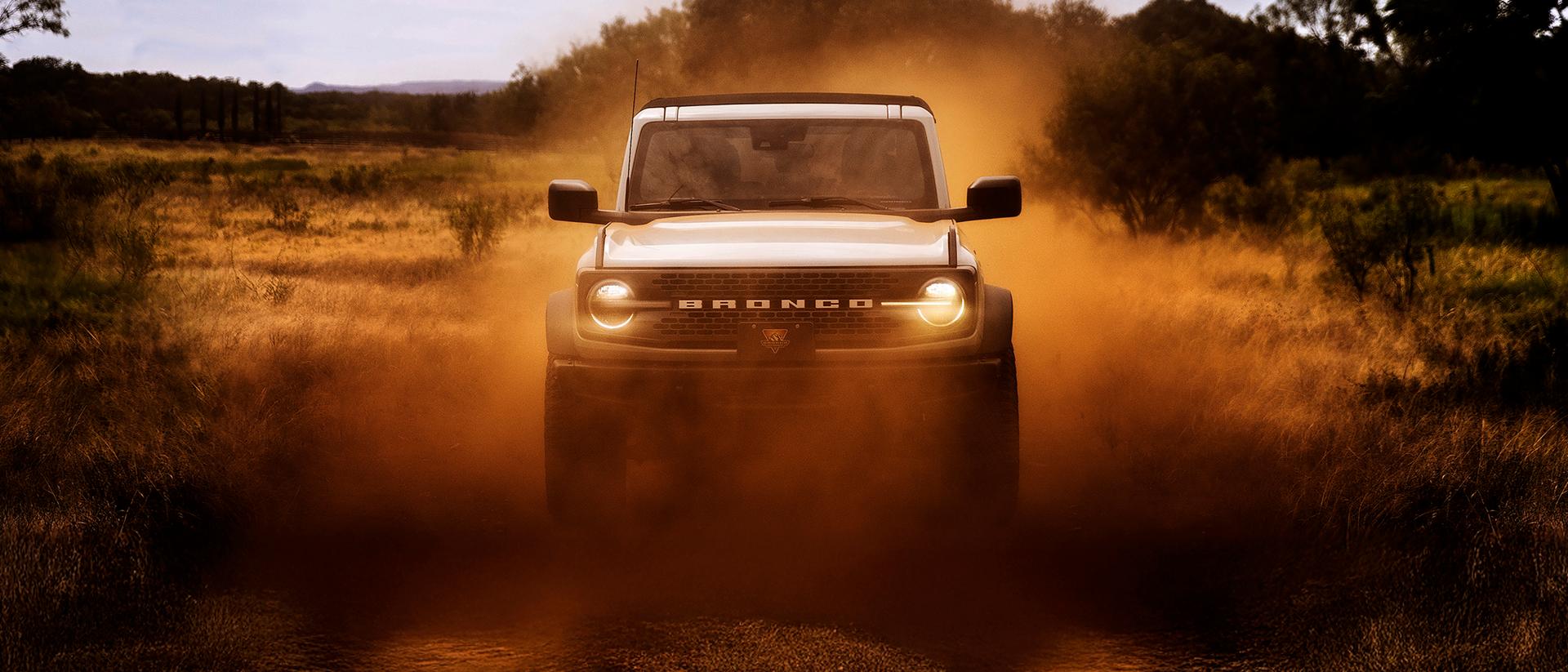 The front of a 2026 Ford Bronco® SUV, its headlights shining through a cloud of red dust