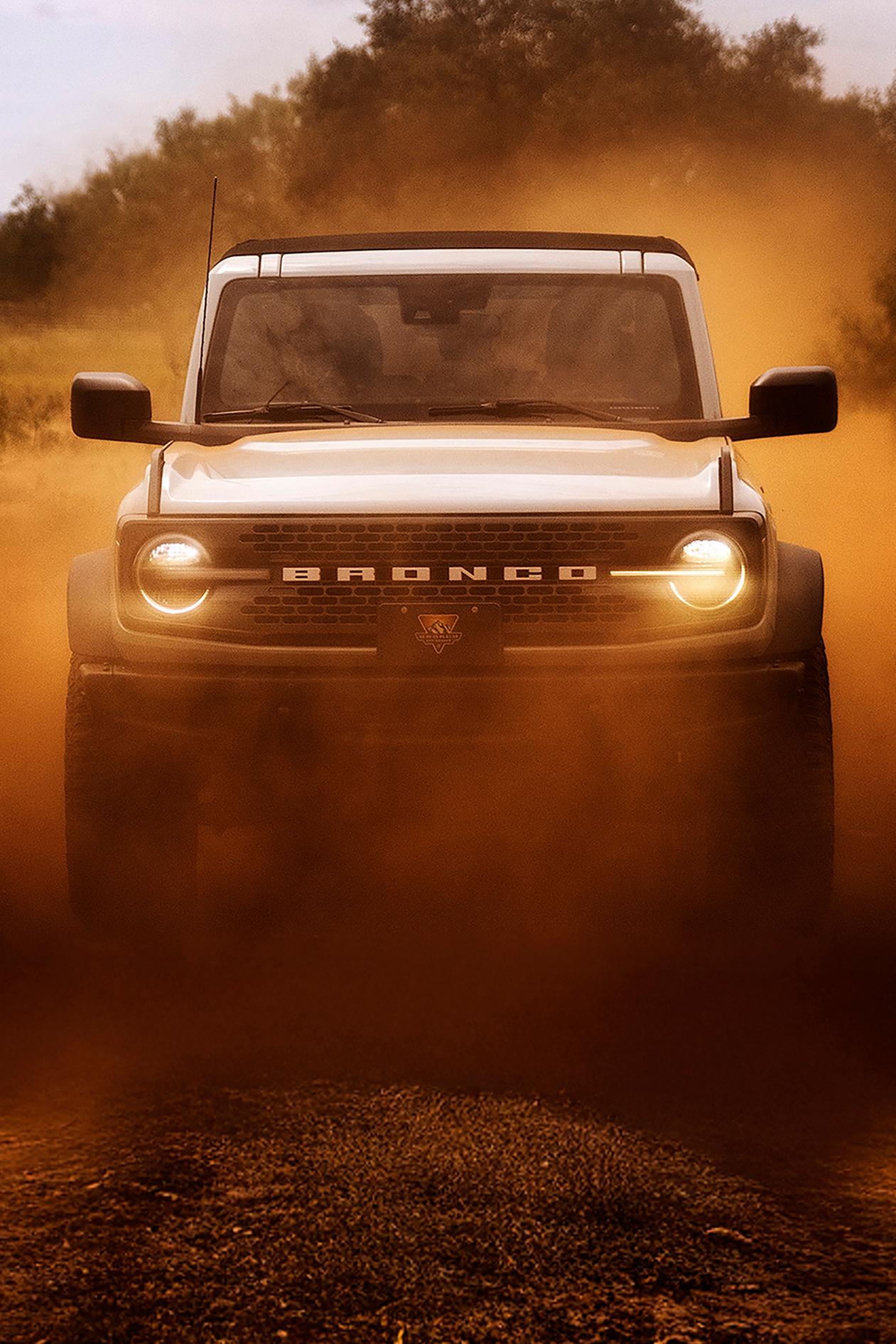 The front of a 2026 Ford Bronco® SUV, its headlights shining through a cloud of red dust