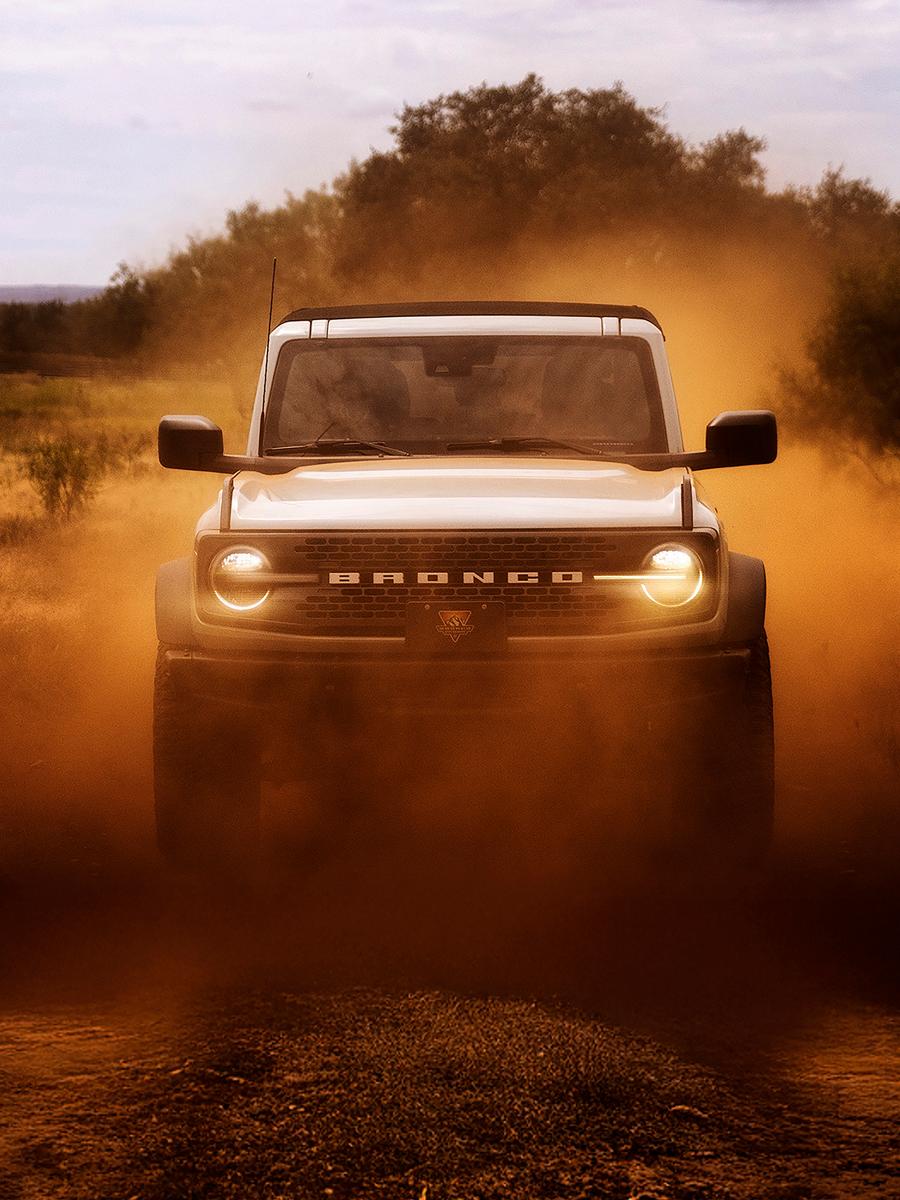 The front of a 2026 Ford Bronco® SUV, its headlights shining through a cloud of red dust