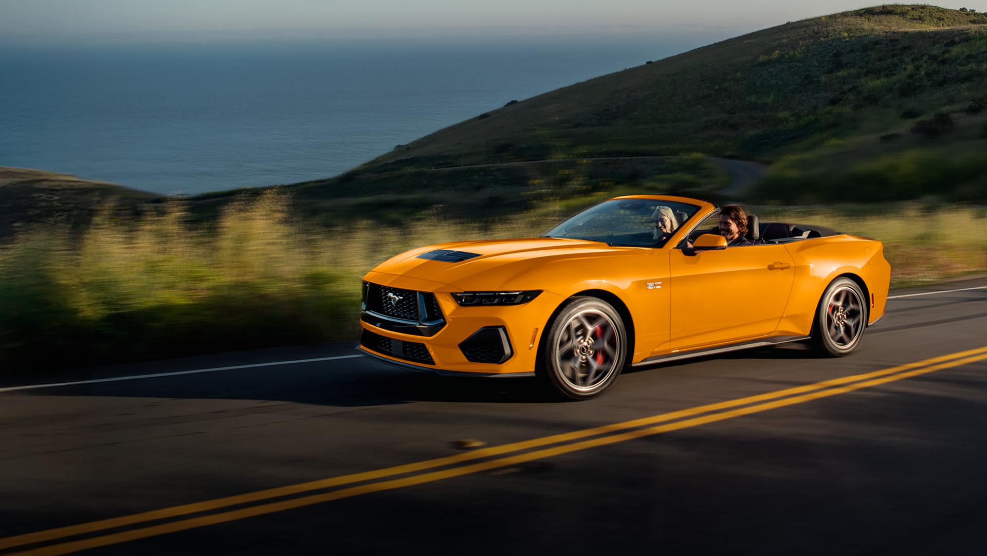 Two people in a 2026 Ford Mustang® GT convertible in Orange Fury Metallic Tri-coat are being driven on a coastal highway