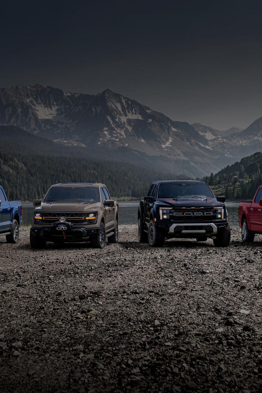 Four 2025 Ford F-150® trucks parked on a rocky road with a lake and mountains in the distance
