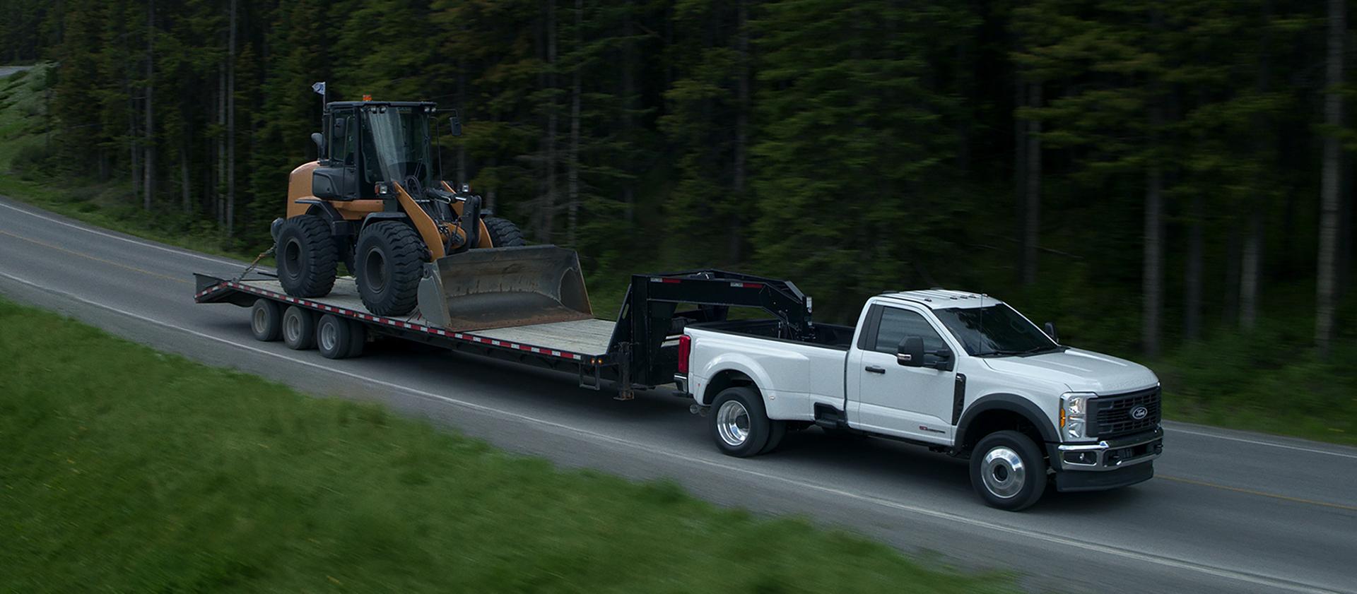 2027 Ford Super Duty® F-450® XL model towing a flatbed trailer carrying a compact wheel loader on a paved road in the mountains