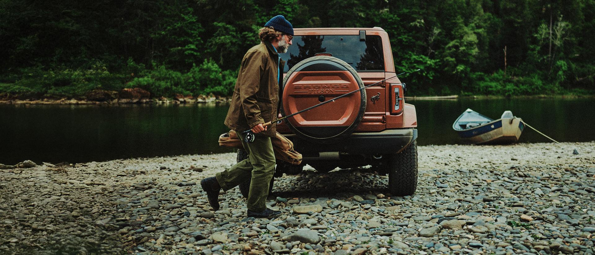 A man carrying a fishing pole and a storage bag near a 2027 Ford Bronco® Filson SUV parked on a rocky lakeshore