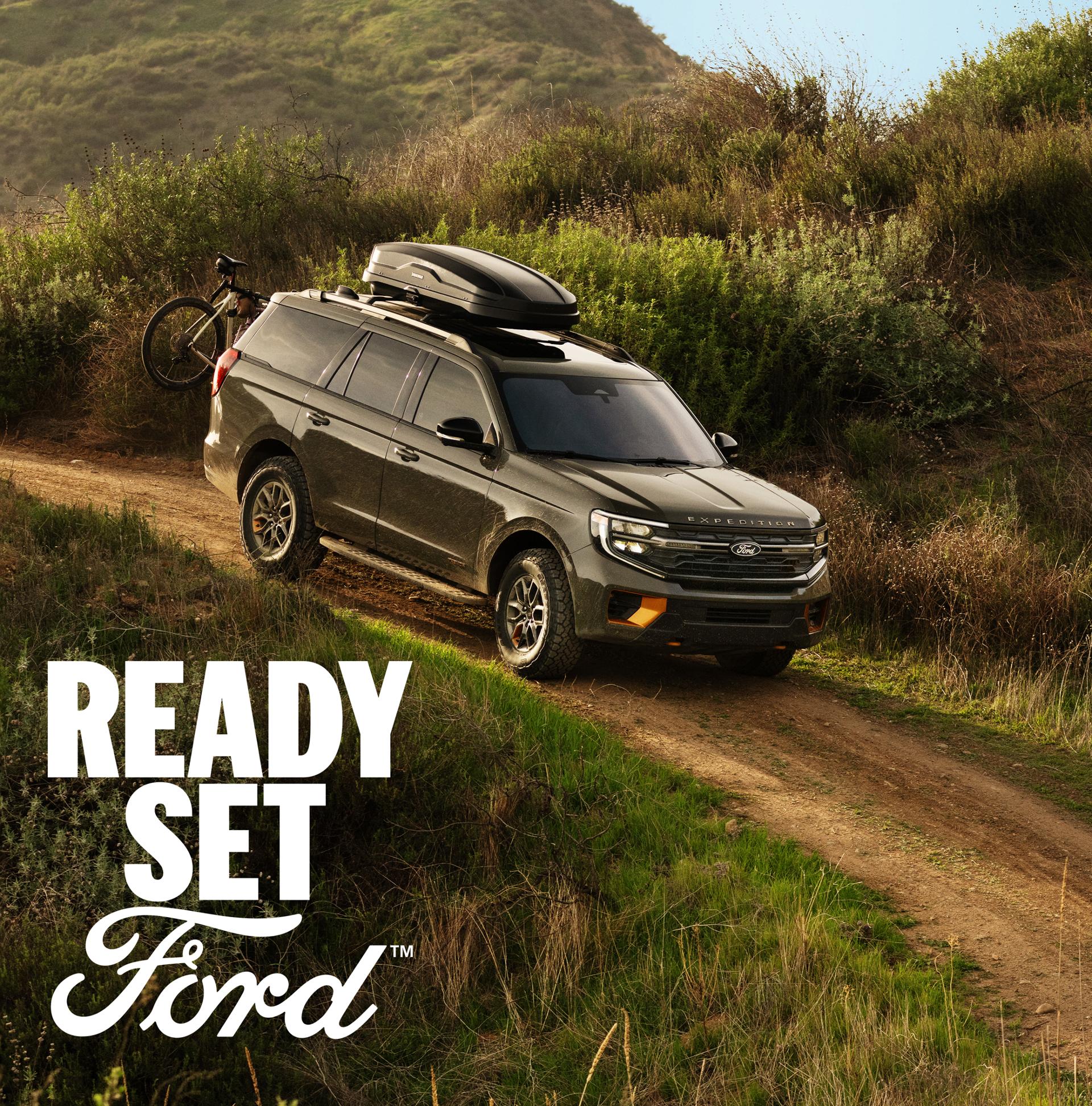 A side profile view of the 2025 Ford Expedition in wild green on a dirt trail with a mountain bike mounted on the back. White text “Ready Set Ford” appears in the lower left-hand corner. 