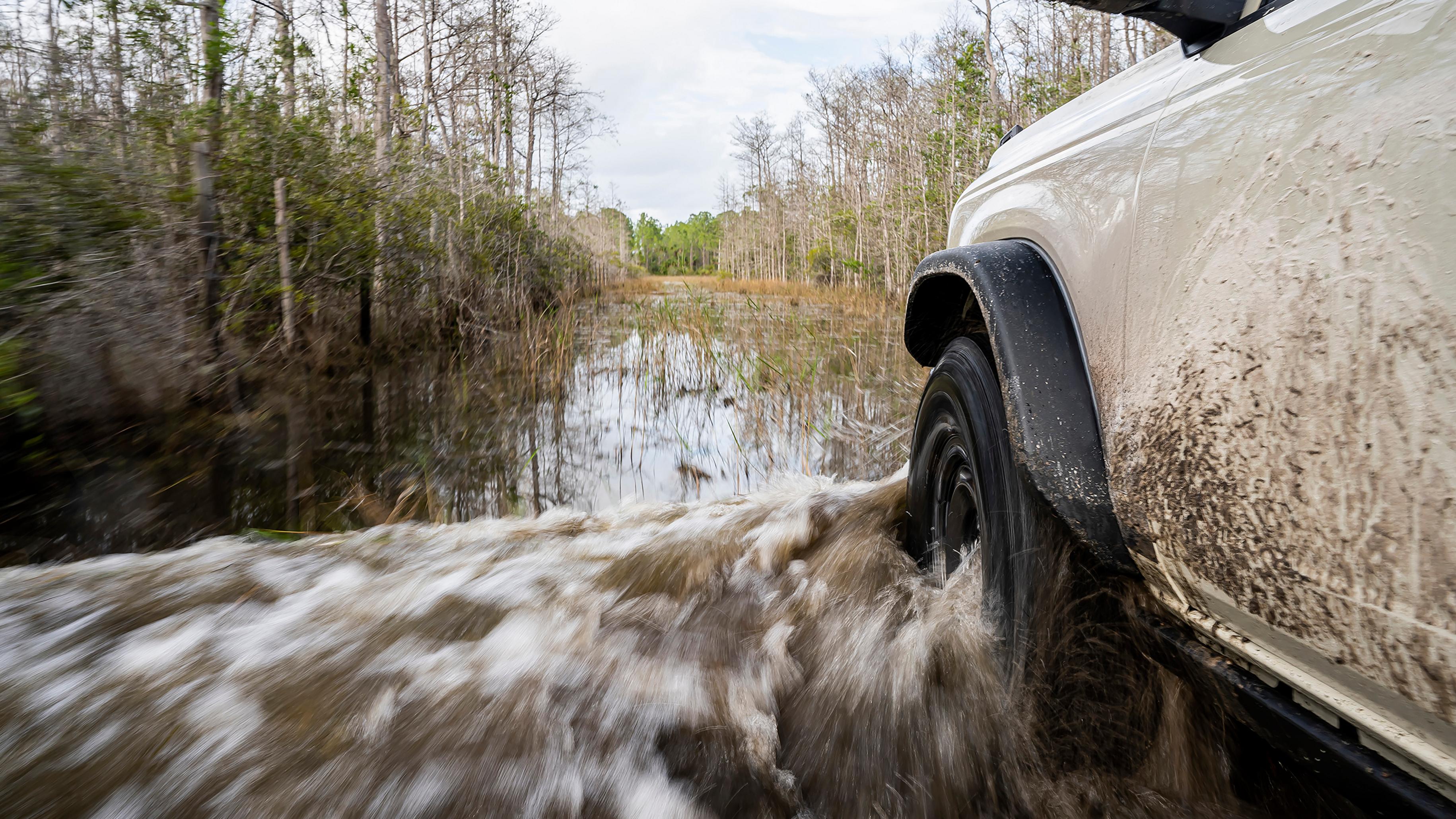 A close-up of the front driver's-side wheel and fender of a 2026 Ford Bronco® SUV as it's driven through a river