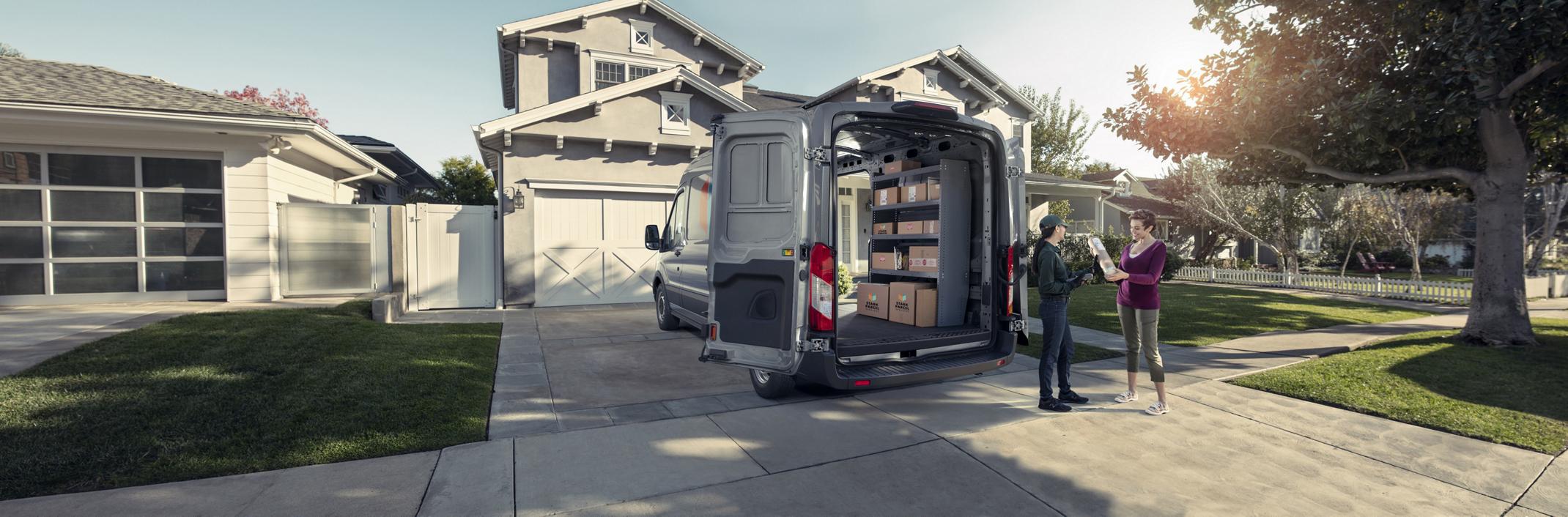 A 2025 Ford Transit® van parked in a driveway with rear doors open as a delivery person drops off a package