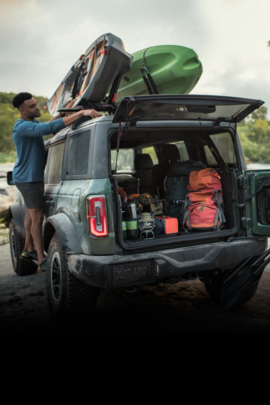 A green 2024 Ford Bronco® Outer Banks® model parked with the tailgate open. A women and man are unloading for a camping trip. Kayaks are strapped to the top.