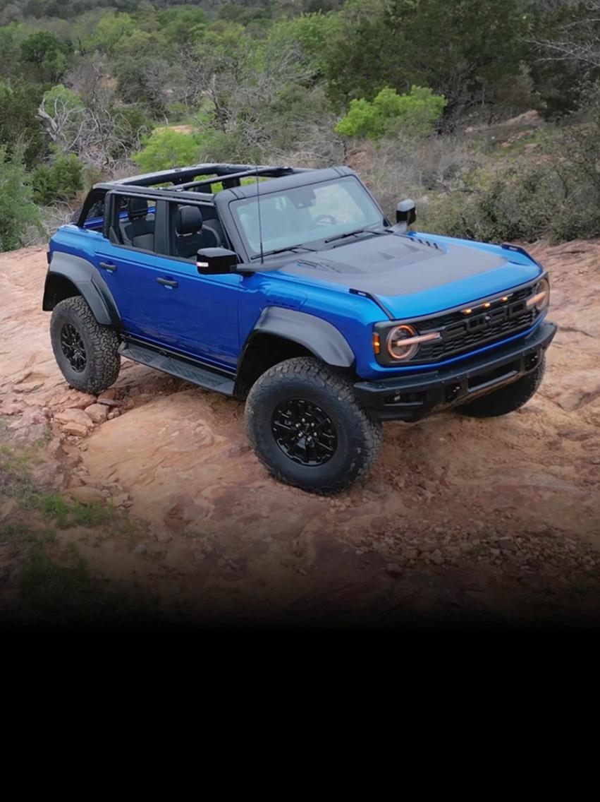 A blue 2026 Ford Bronco® Raptor® with its top off is parked on a dirt hill in a rugged, brush-filled environment