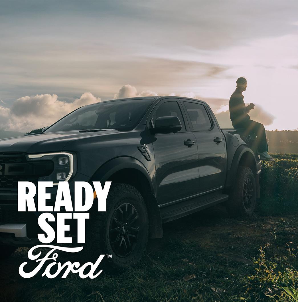 Three-quarter view of a grey Ford Ranger Raptor parked on grass. A person sits on the end of the truck bed, the sun shining in the background. White text “Ready Set Ford” appears in the lower-left corner.