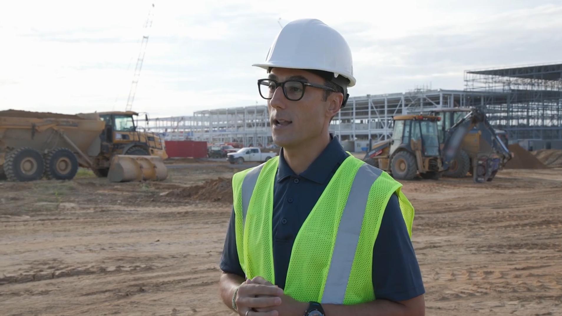 Standing in front of an active construction site, a person in a hard hat and reflective vest speaks to someone off camera