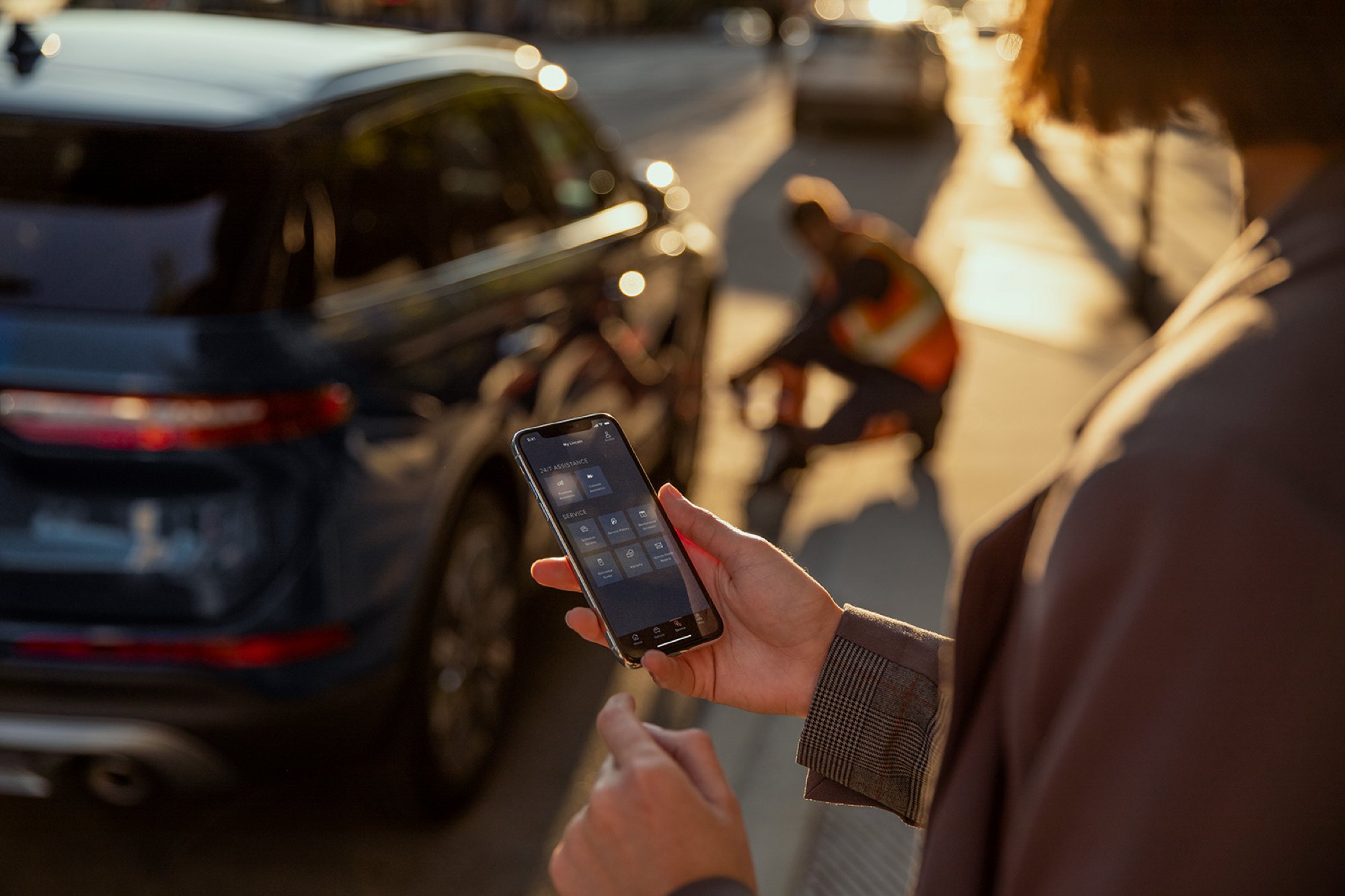 A woman is shown looking at her mobile device while someone changes a tire on a nearby Lincoln vehicle.