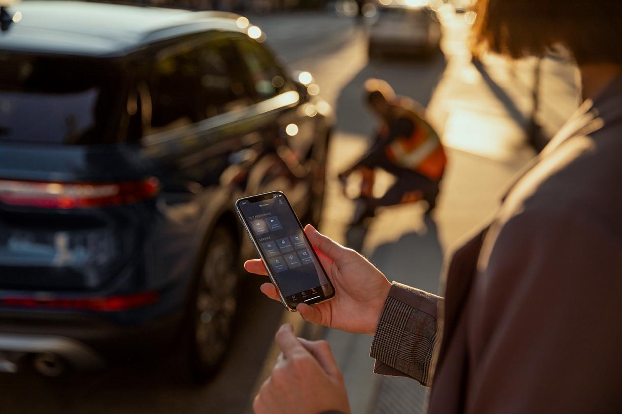 A woman is shown looking at her mobile device while someone changes a tire on a nearby Lincoln vehicle.