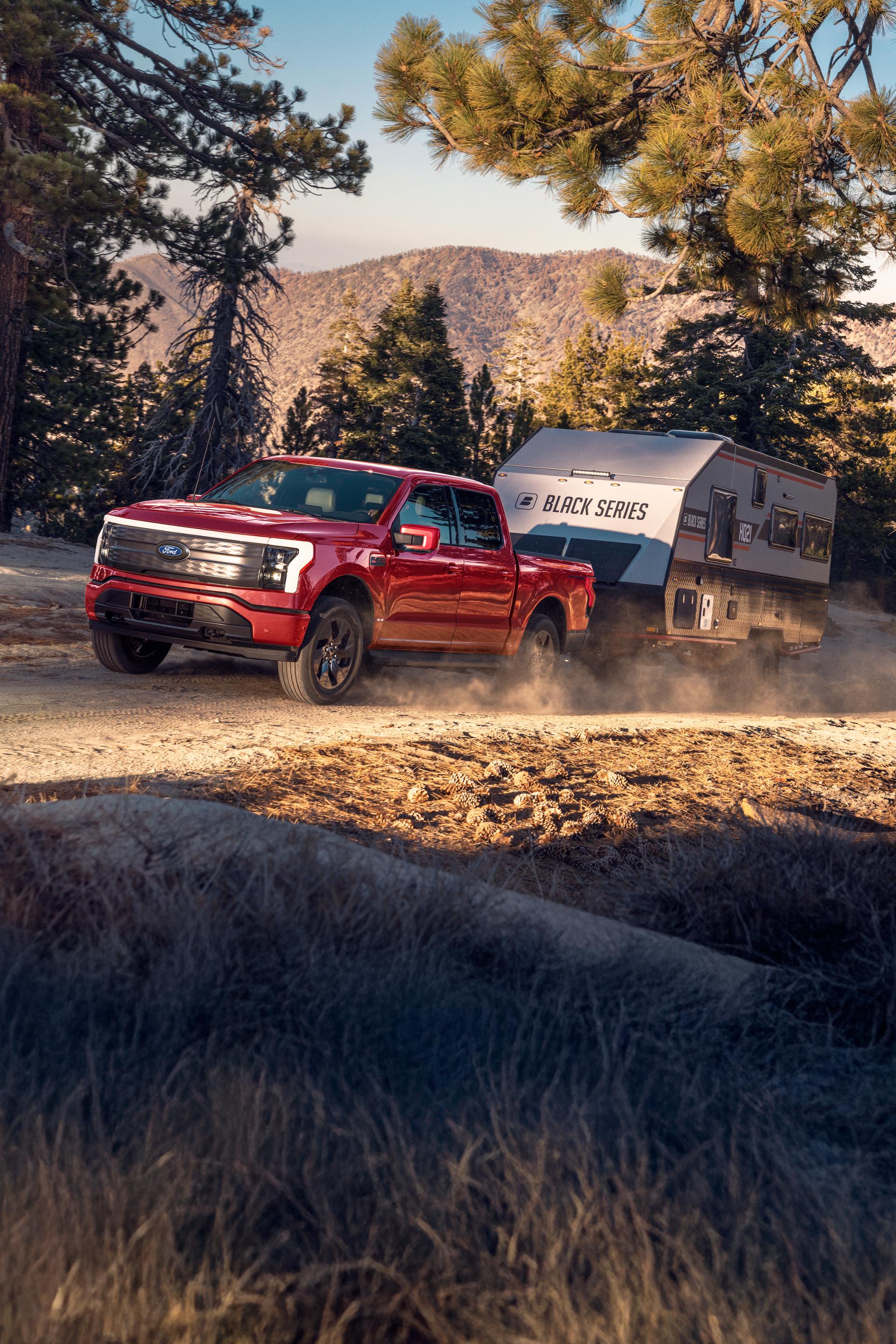 A red 2025 Ford F-150® Lightning® truck towing a trailer being driven down a mountainous and forested road