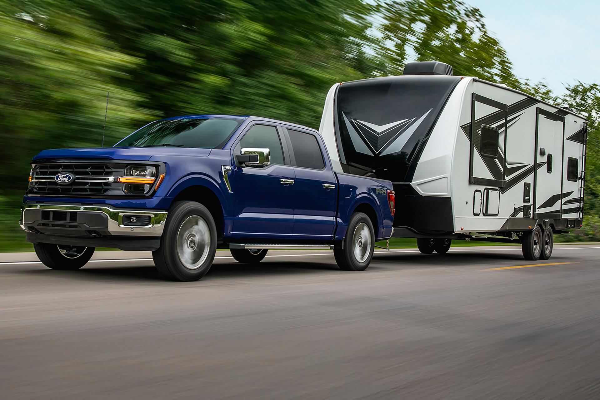 A blue Ford F-150® truck towing a camper down a forest-lined highway