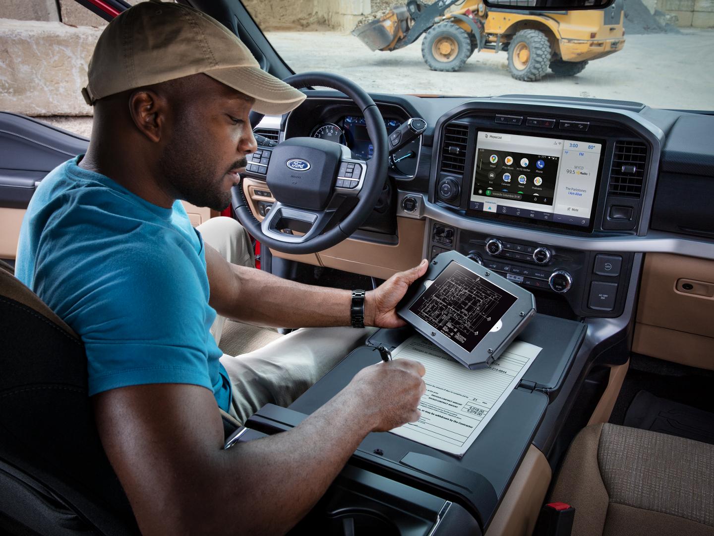 A man in the driver's seat of a 2022 Ford F-150 parked at a construction site; a front loader is visible through the windshield. The man is holding a tablet computer with his left hand and writing on a paper with his right hand. The vehicle's SYNC touchscreen is displaying a menu of icons.