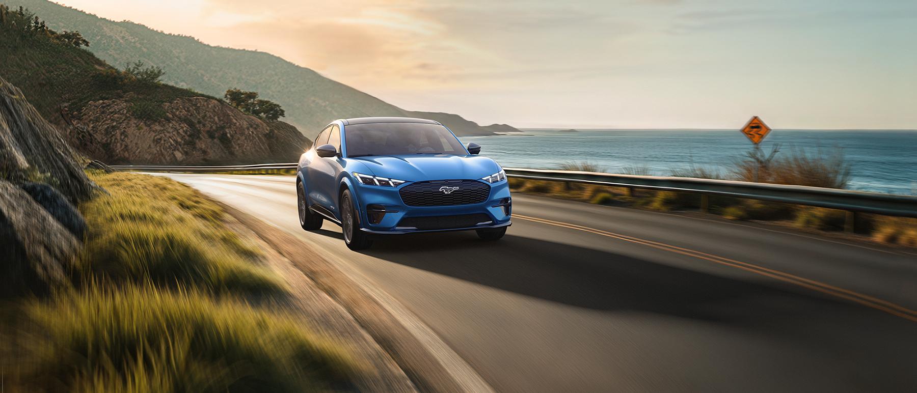 The Ford Mustang Mach-E® SUV is driven around a bend on a coastal highway at sunset